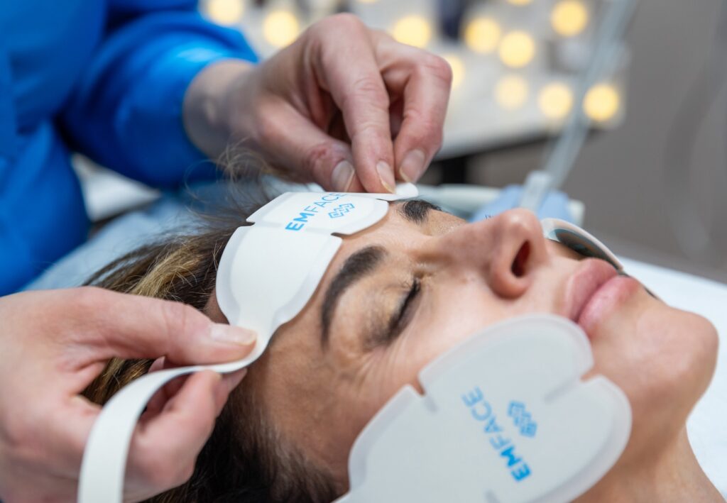 Patient lying back with Emface applicators on her cheeks while the provider places applicators on her forehead. This is an appointment for facial contouring in Jacksonville, NC.