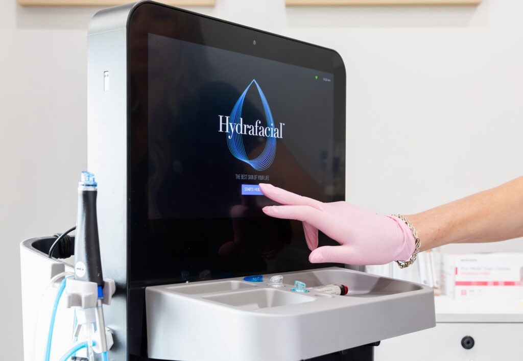 A gloved medical provider selects settings on a HydraFacial touchscreen in a treatment room. The handheld device is placed on the side of the computer screen as the provider prepares for a customized facial in Jacksonville, NC.
