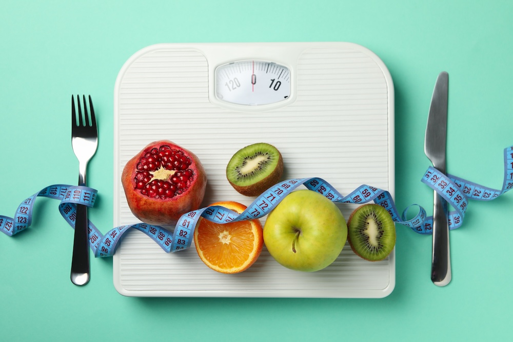 A bathroom scale on a blue background with some fruit across it, a fork on the left, a knife on the write, and a blue body measuring tape winding across the top. This shows the importance of realistic goals and healthy habits when working with a weight loss clinic in Jacksonville.