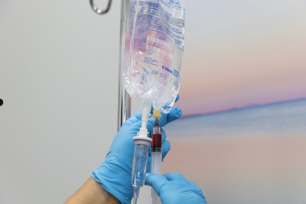 Provider wearing gloves connects tubes to an IV drip in Jacksonville, NC. The drip bag is hanging from a pole and contains a mix of vitamins and minerals to support patient's immunity.